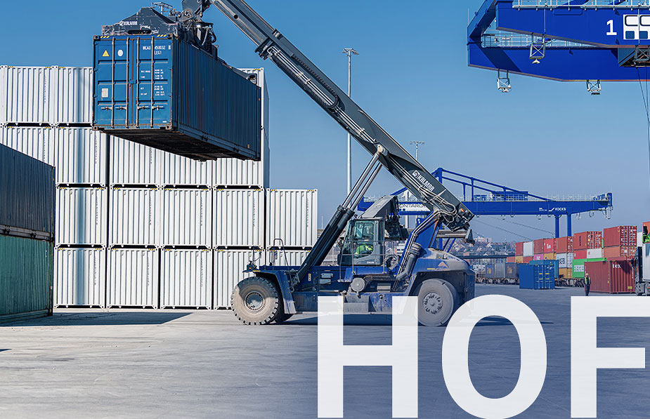 Blauer Reachstaker, der einen blauen Container transportier. Im Hintergrund ein Container Terminal mit einem Kran.