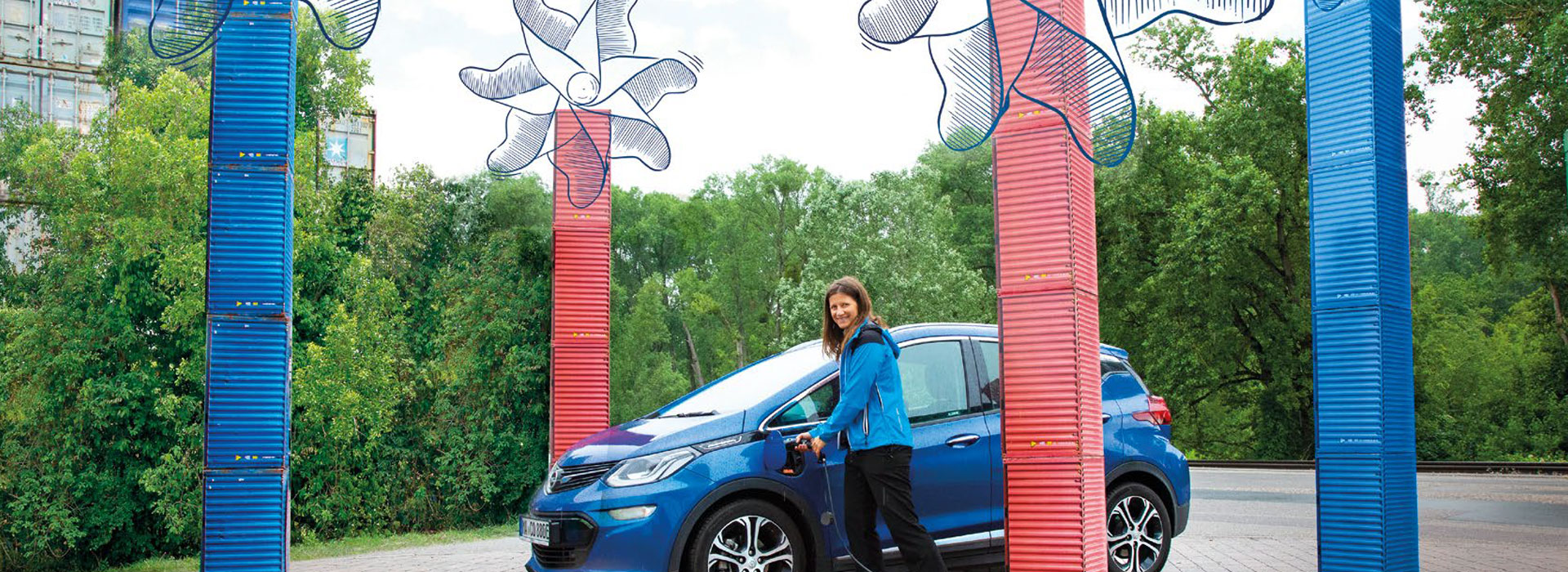 E-Auto mit Ladekabel inmitten von Pappcontainern, die wie Windräder aussehen