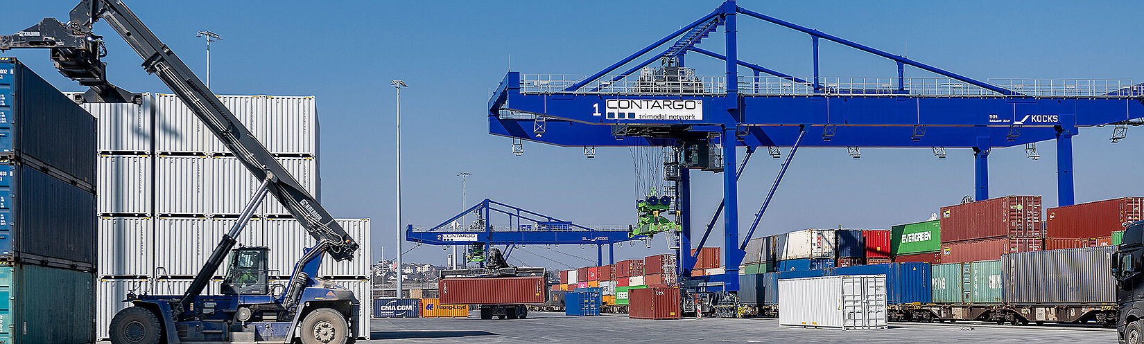 Links hebt ein Reachstacker einen Container an. Rechts sind zwei blaue Krananlagen mit diversen Containerstapeln