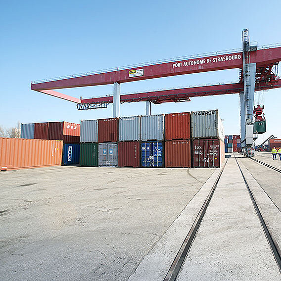 Container Terminal mit rotem Kran. Gestapelte Container und Bahnschienen darunter.