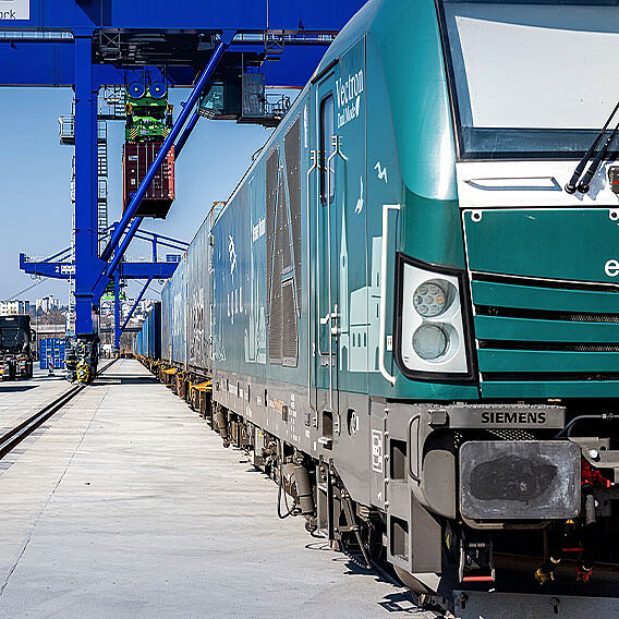 Dunkelgrüner Container Zug. Lok im Vordergrund. Container Terminal und Kran im Hintergrund.