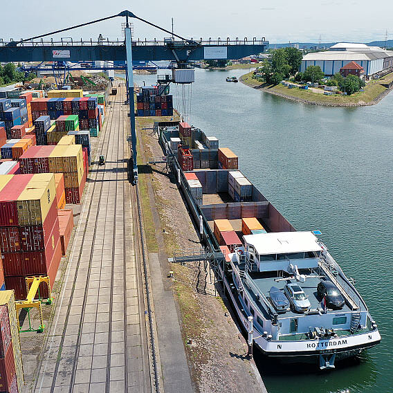 Container Terminal am Fluss mit einem blauen Kran. Container wird auf ein Binnenschiff verladen.