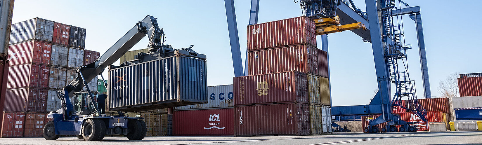 Reachstacker mit Container beladen fährt über Container-Terminal mit Krananlage im Hintergrund