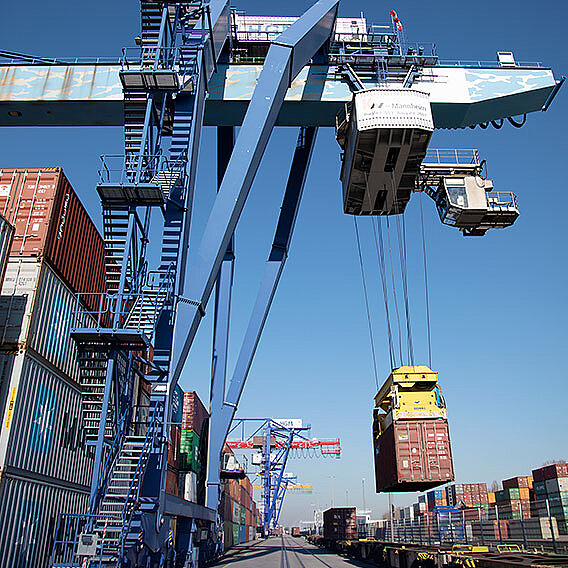 Container Terminal mit einem großen blauen Kran im Vordergrund, der Container verlädt.