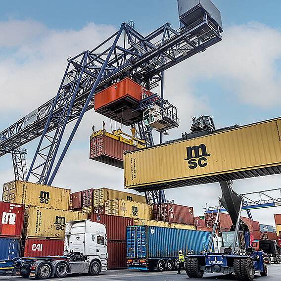 Reachstacker mit gelbem Container. Im Hintergrund LKW, Container und ein blauer Kran