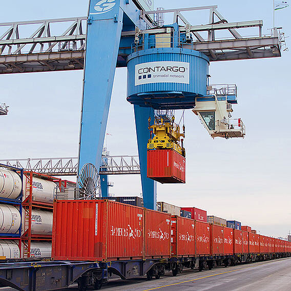 Blauer Kran, der rote Container auf einen Container Zug verlädt.